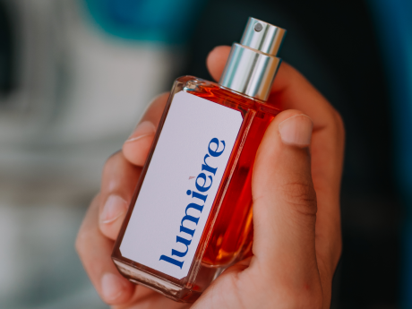 Hand holding a red bottle of Lumiere perfume against a blurred background